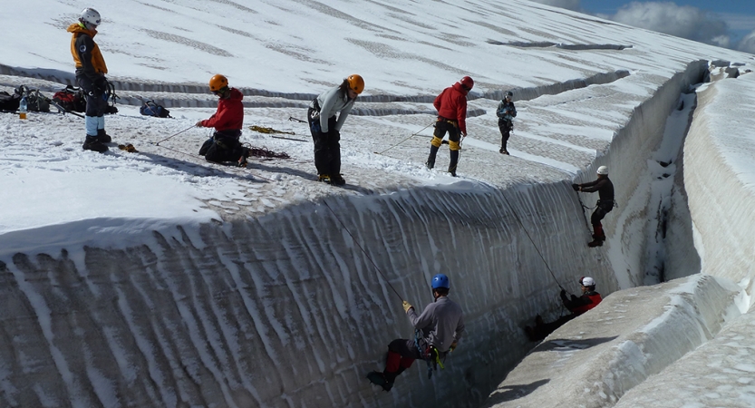 A group of people wearing safety gear are secured by ropes as they rappel down a snowy bank. Others stand on the top of the bank.